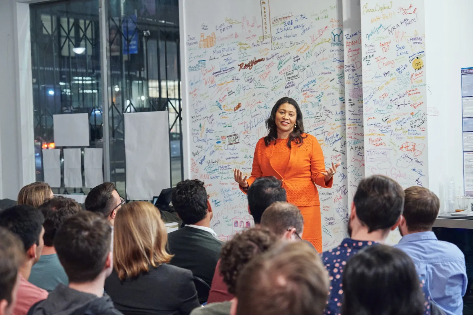 A speaker addressing a full room in front of a wall covered in members’ signatures
